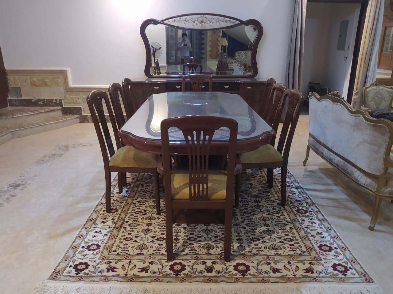 طاولة طعام وكراسي خشبية وخلفها خزانة بمرآة، فوق سجادة كريمية زهرية